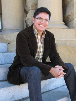 Juan sitting outside on steps in jeans, a light plaid shirt with brown jacket.